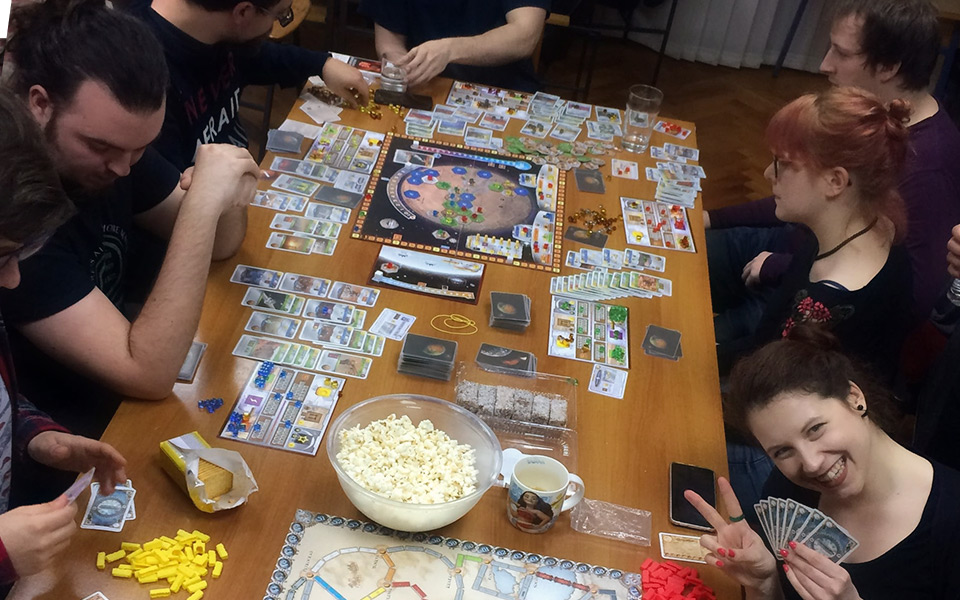 Više osoba koje za stolom igraju društvene igre, uz razne grickalice.