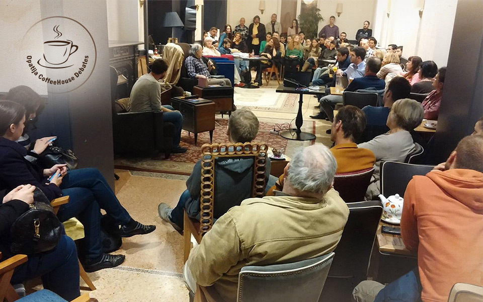 Velika grupa ljudi koja sjedi u Kavani Hotela Continental u Opatiji, slušaju gostujućeg predavača. U gornjem lijevom kutu nalazi se logo Opatija Coffeehouse Debates sa šalicom kave unutar tankog kruga tamnosmeđe boje.