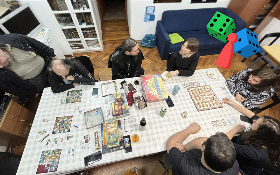 Sedmero ljudi okupljenih za stolom na kojemu se nalaze društvene igre, u prostorijama udruge Kulturni front. U gornjem desnom kutu nalazi se šarena ilustracija s dvjema kockicama i pijunom.