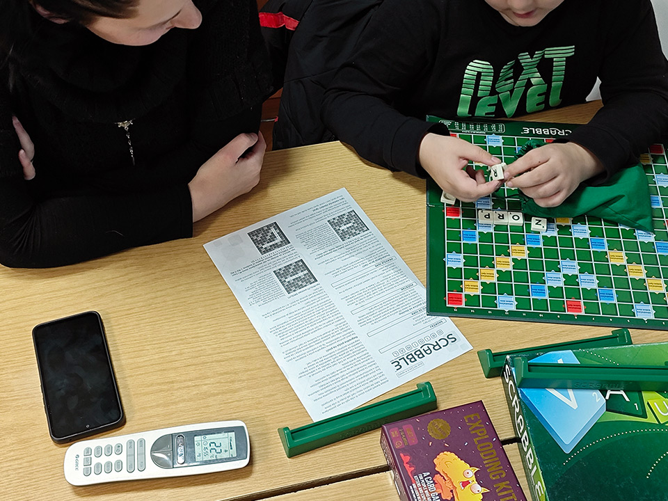 Žena i dijete koji igraju Scrabble za stolom. Fokus je na upute i zelenu ploču igre.