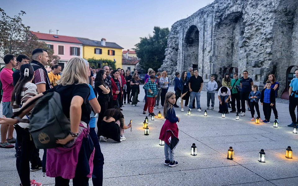 Poveća grupa ljudi, okupljena oko lampiona koji su kružno postavljeni po kamenom podu.